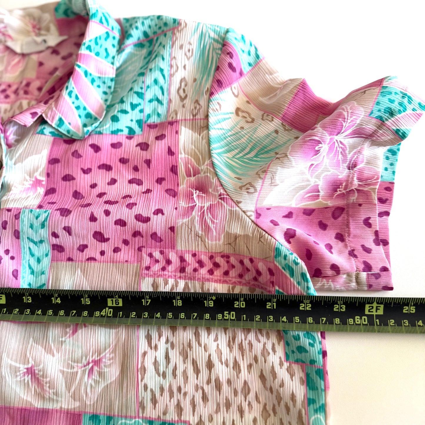 Button down bright coloured (tan, pink, teal) shirt with tropical and animal print patterns on it - placed on a white background with a measuring tape on it.