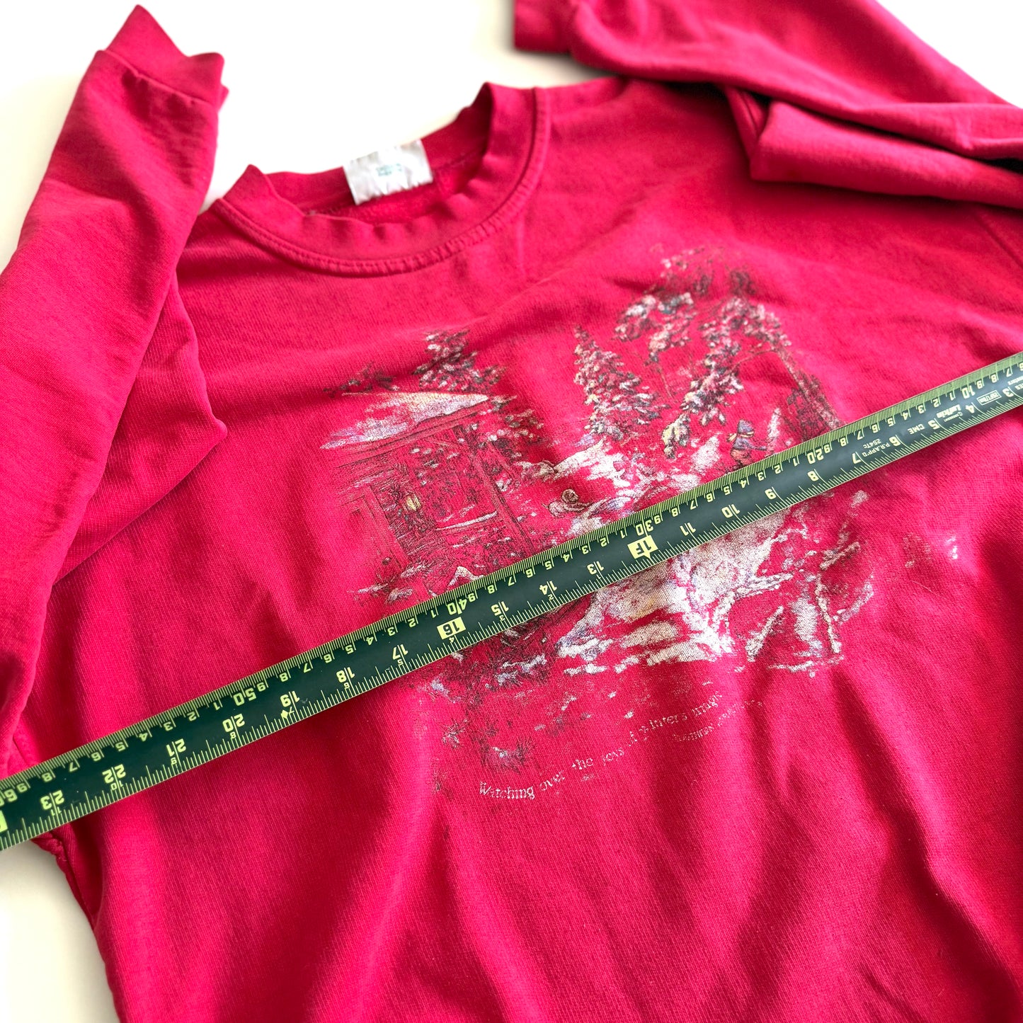 Bold pink sweatshirt / crewneck with a winter scene on the front laid on a white backdrop - with a measuring tape laid out on it.