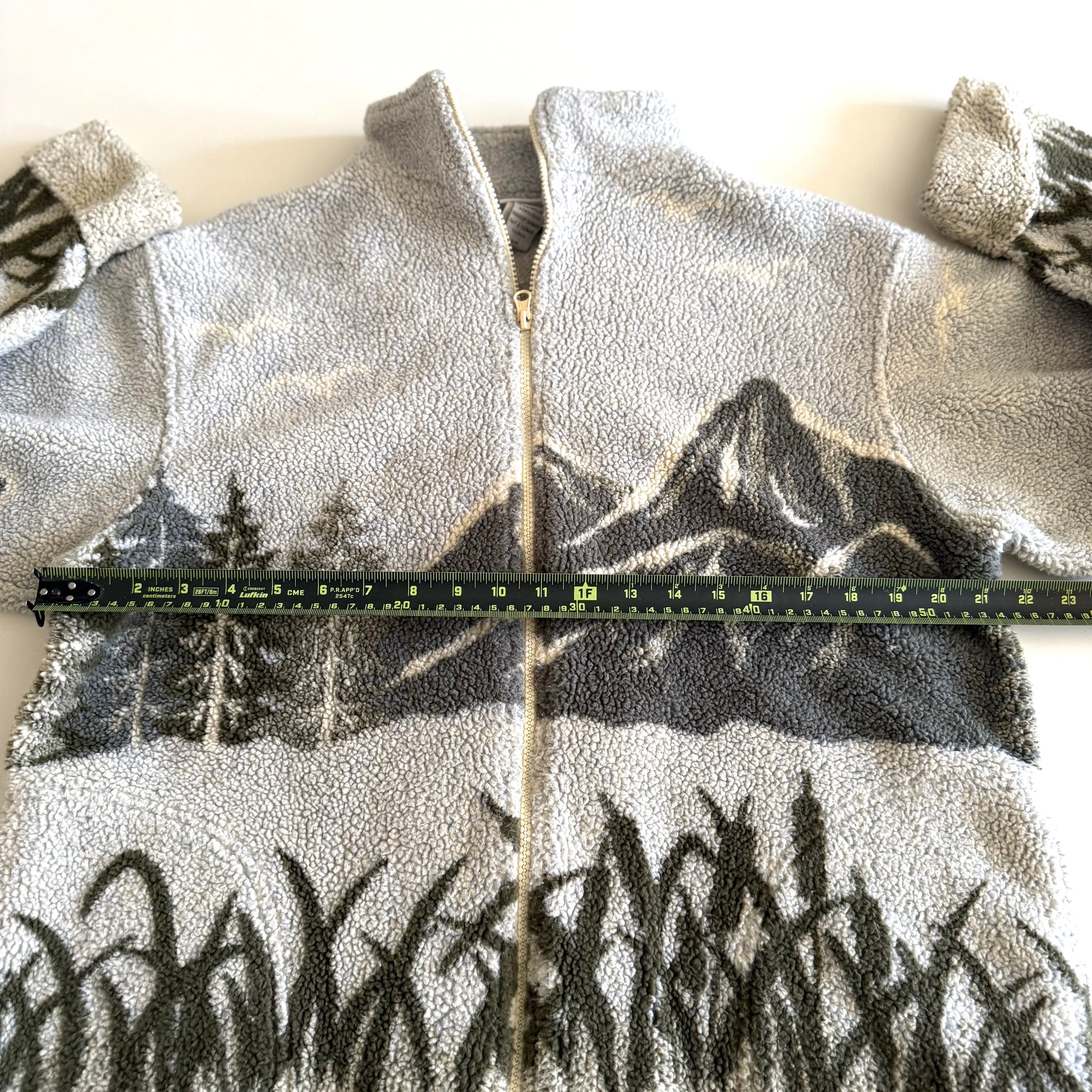Fleece textured sweater with a mountain marsh forest scene on it, and a zippered close - on a white backdrop with a measuring tape laid  on it. 