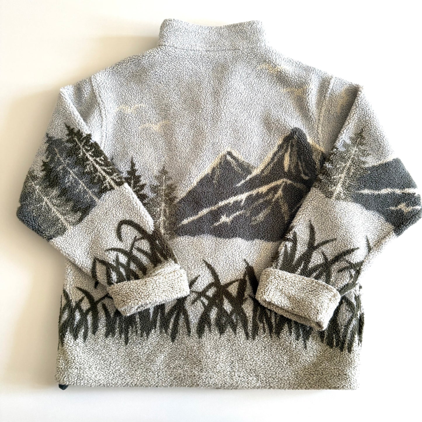 Backside of fleece textured sweater with a mountain marsh forest scene on it, and a zippered close - on a white backdrop with a measuring tape laid  on it. 